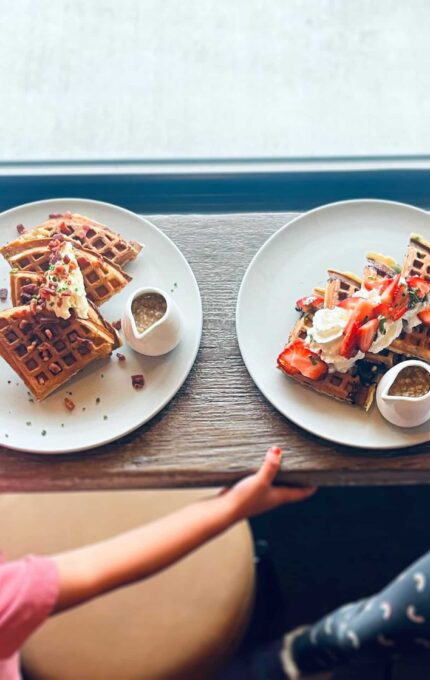 Two children are seated at a counter with plates of waffles in front of them. Each plate features waffles topped with various ingredients, and small containers of syrup are placed alongside. The children are partially visible, with one reaching towards the other.