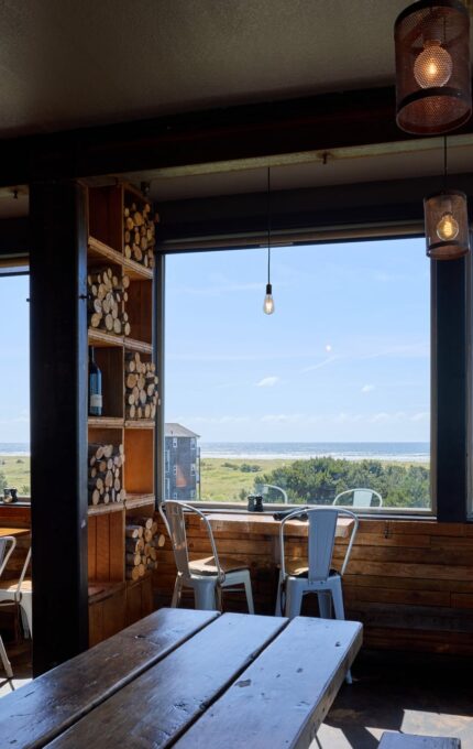 A small, cozy cafe with wooden tables and metal chairs is shown. Large windows offer a scenic view of a beach and ocean in the background. Stacks of firewood are seen in a corner shelf, and pendant lights hang from the ceiling.