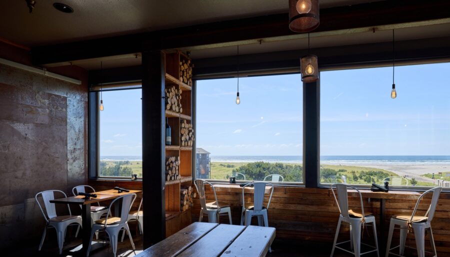 A small, cozy cafe with wooden tables and metal chairs is shown. Large windows offer a scenic view of a beach and ocean in the background. Stacks of firewood are seen in a corner shelf, and pendant lights hang from the ceiling.