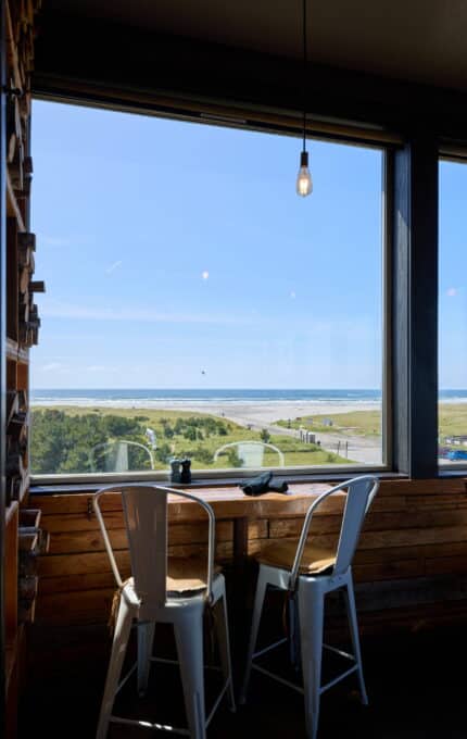 A coastal landscape is visible through a large window in a room. Two white metal chairs are positioned at a wooden table in front of the window. A single lightbulb hangs from the ceiling. The view outside includes grassy dunes, sandy beach, and blue sky.