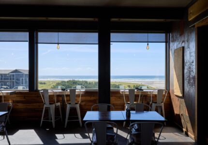 Interior of a cafe with tables and chairs positioned next to large windows that offer a scenic view of the ocean and surrounding landscape. Ceiling lights hang above the tables, and the wooden decor adds a rustic touch to the setting.