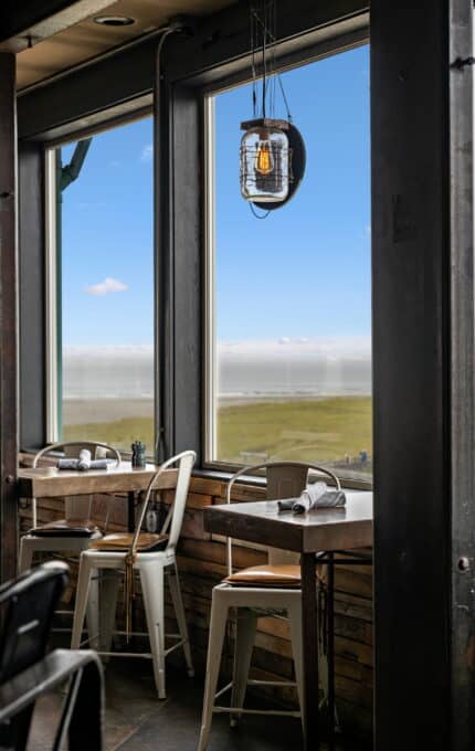 A small dining area with wooden tables and white metal chairs is adjacent to large windows offering a scenic view of a grassy field and blue sky. A rustic light fixture hangs from the ceiling. Napkins and utensils are neatly placed on the tables.