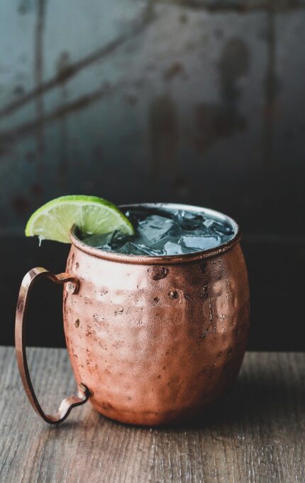 A copper mug filled with a dark beverage topped with ice and a lime wedge on the rim sits on a wooden surface. The background is blurred, featuring a textured, rustic wall.