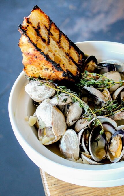 A white bowl filled with steamed clams garnished with fresh herbs and accompanied by two pieces of grilled bread is placed on a wooden surface. The background is blurred, showcasing a rustic, textured floor.