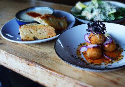 Three plates of food on a wooden table. The first plate has fried balls topped with red onions and sauce. The second plate contains slices of bread with a dipping sauce. The third plate has a green salad with grated cheese and slices of avocado.