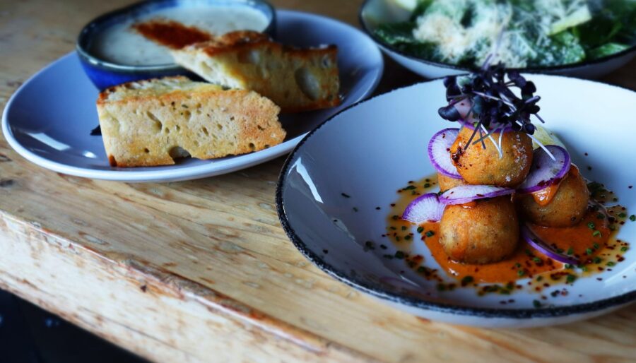 Three plates of food on a wooden table. The first plate has fried balls topped with red onions and sauce. The second plate contains slices of bread with a dipping sauce. The third plate has a green salad with grated cheese and slices of avocado.