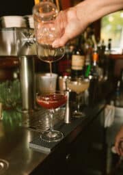 A hand is shown pouring liquid from a glass shaker into a coupe glass containing a dark red cocktail. Another coupe glass with a pale yellow drink is in the background, along with various bottles and bar equipment on a countertop.