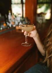 A person with long hair is holding a coupe glass filled with a light-colored cocktail at a wooden bar. The background is blurred, featuring various bottles and bar equipment.