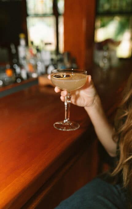 A person with long hair is holding a coupe glass filled with a light-colored cocktail at a wooden bar. The background is blurred, featuring various bottles and bar equipment.