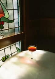 A cocktail in a coupe glass sits on a round white marble table near a stained glass window. The sunlight filters through the window, casting soft shadows on the table.