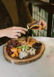 A person spreads jam on a piece of toast from a wooden platter containing grapes, sliced green apples, cubes of cheese, a clump of cheese, and a small bowl of jam. The scene is set at a table with a stained glass window in the background.