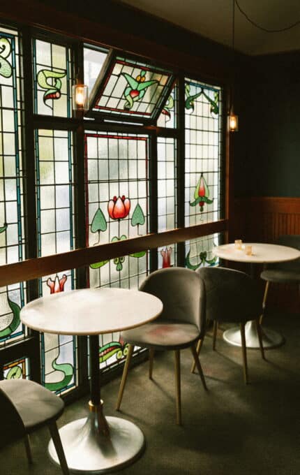 A small, cozy seating area in a cafe featuring round white tables, gray chairs, and decorative stained glass windows with floral motifs and green borders. The lighting is soft, with small pendant lights hanging from the ceiling.