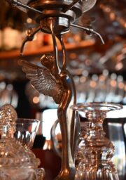 Close-up of a bar shelf with various crystal glassware and a metallic sculpture of an angel with wings. The background shows blurred bottle tops and hanging glass goblets. The scene is warmly lit, highlighting the reflective surfaces of the glassware and sculpture.