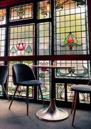 A small seating area with two black chairs and a round metal table is set against a backdrop of stained glass windows. The windows feature decorative patterns and floral designs, letting in natural light and creating a colorful ambiance.