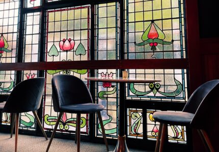 A small seating area with two black chairs and a round metal table is set against a backdrop of stained glass windows. The windows feature decorative patterns and floral designs, letting in natural light and creating a colorful ambiance.