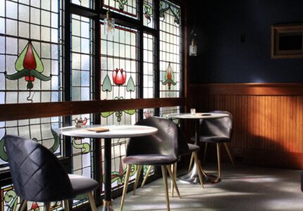 A small café interior with stained glass windows, two round white tables, and dark gray chairs. The room has wooden paneling on one wall, soft lighting, and a cozy ambiance. An unoccupied intimate setting with sunlight streaming through the windows.