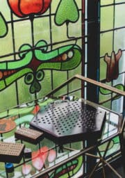 A table with a glass top holds a hexagonal wooden board game and two rectangular wooden pieces with multiple holes. The background features a decorative stained glass window with green, red, and brown patterns.