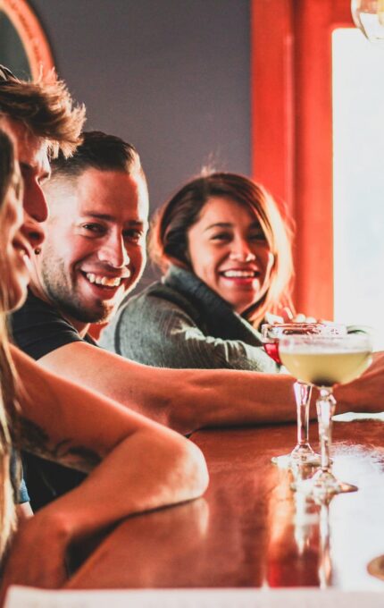 Four people are sitting at a wooden bar counter, smiling and engaging in conversation. They have drinks in front of them, including a margarita and a beer. The bar has dark walls and warm lighting.