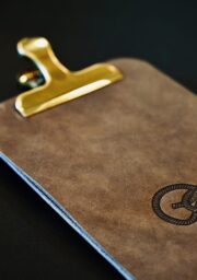 A brown leather clipboard with a golden clip. The surface of the clipboard features a printed or embossed knot design. The background is black.