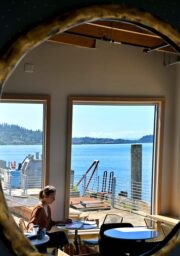 A round mirror with a golden frame reflects a café interior with large windows overlooking a body of water and distant hills. A person is seated at a table, working on a laptop. Bright natural light illuminates the scene.