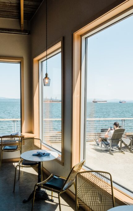 A cafe features metal wire chairs and round tables placed near large windows overlooking a body of water. Outside, a person sits on a lounge chair by the waterfront. Several ships are visible in the distance under a clear blue sky.