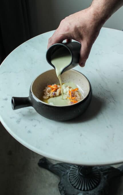 A hand is pouring a light green sauce from a black pitcher into a round dish that contains food, including orange and white elements. The dish is placed on a white marble table. The base of the table is visible in the bottom part of the image.