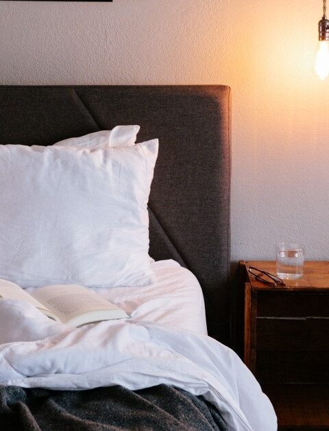 Two made beds with gray headboards are separated by a wooden nightstand holding a glass of water. One bed has an open book and a cushion with a sun and waves pattern. An exposed bulb hangs above the nightstand.