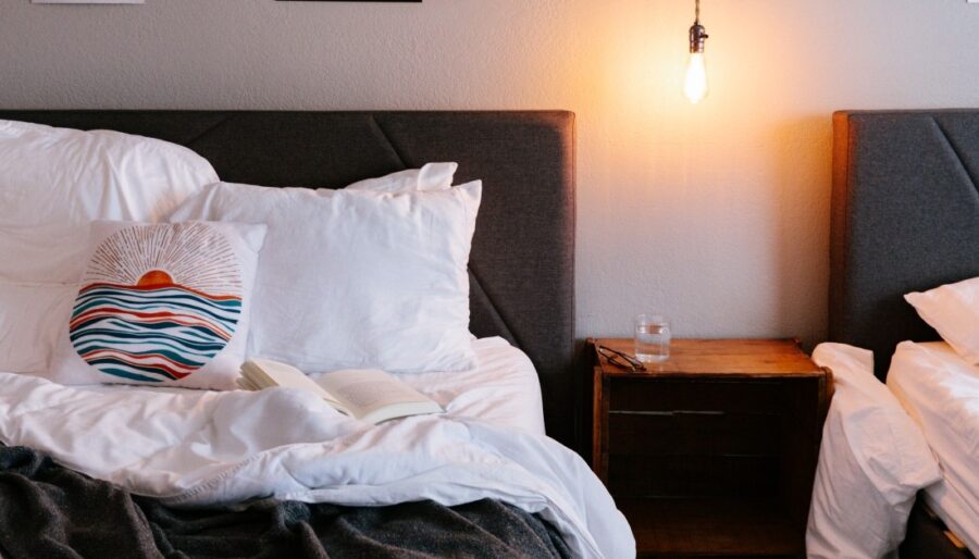 Two made beds with gray headboards are separated by a wooden nightstand holding a glass of water. One bed has an open book and a cushion with a sun and waves pattern. An exposed bulb hangs above the nightstand.
