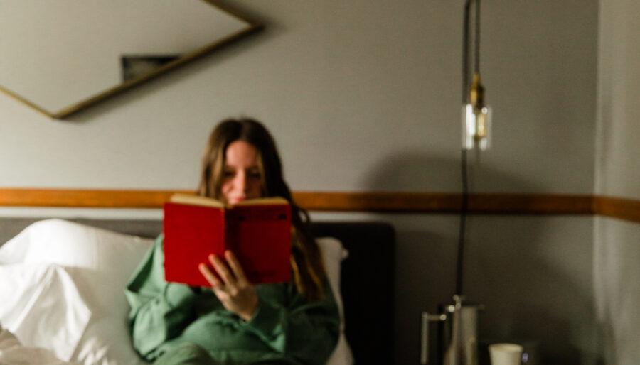 A person in green loungewear sits on a bed reading a red book. Their bare feet are visible in the foreground. A bedside table with a carafe and glass sits nearby, and a geometric mirror hangs on the wall.