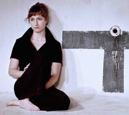 Woman sitting on the floor against a white wall with a large black "T" shape painted on it. She is wearing black clothing, has short hair, and is looking slightly off to the side. The floor and wall have a rough, unfinished texture.