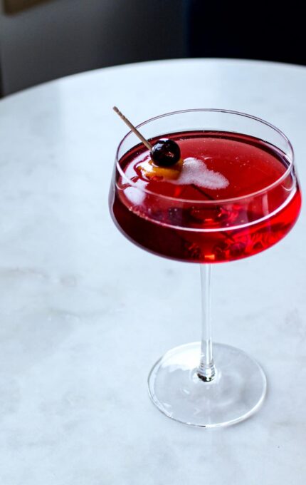 A clear stemmed cocktail glass filled with a red drink garnished with a single cherry on a wooden stick sits on a white marble surface. The table is slightly reflective, and a part of a grey wall is visible in the background.