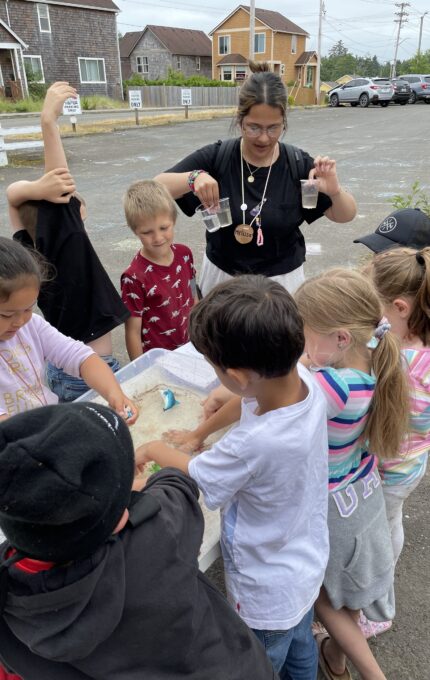 A group of children and an instructor gather around a table outdoors. The children are engaging in a hands-on activity involving sand. The instructor is holding two jars, demonstrating something to the group. Buildings and parked cars are visible in the background.