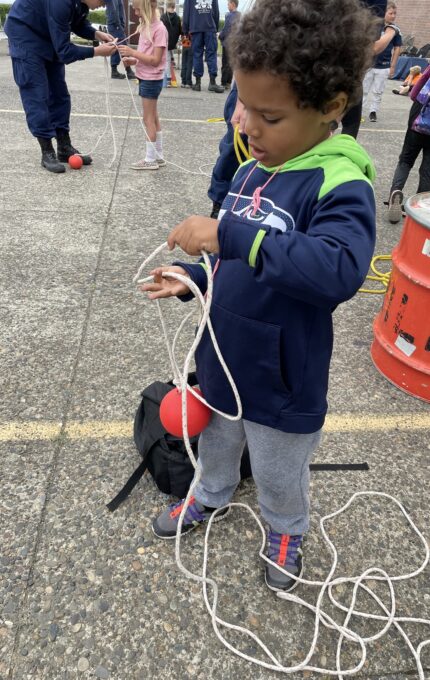 A young child stands on a paved surface holding a long white rope and is seemingly focused on it. The child is wearing a navy blue and green hooded jacket, gray pants, and colorful shoes. Other people and various objects are visible in the background.