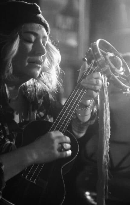 A black and white photo of a person playing a ukulele and singing into a vintage-style microphone. The person is wearing a beanie and bracelets, with eyes closed, creating a focused atmosphere.