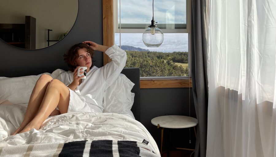 A person in a white dressing gown sits on a bed, holding a mug, next to a window with sunlight streaming in. The room features dark walls, a round mirror, and a blanket with black and white stripes.
