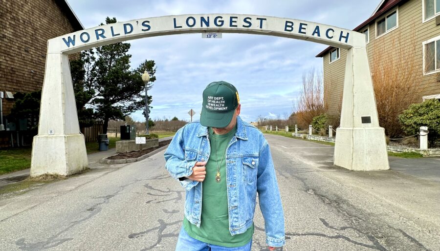 A person wearing a green cap, green shirt, denim jacket, and blue jeans stands on a road beneath an archway that reads "World's Longest Beach." The sky is cloudy, and houses are visible on either side of the road.