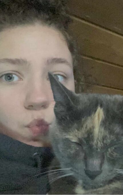 A person with curly hair wearing earbuds is taking a selfie with a cat. The person has a neutral expression with puckered lips, and the cat, which has a mix of gray and orange fur, appears to be relaxed with its eyes closed.