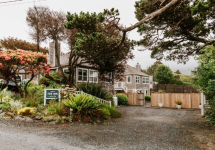 A gravel driveway leads to a rustic inn surrounded by lush greenery and trees. The inn has a stone exterior with large windows. A sign near the entrance reads "The Inn." Flowering plants and a wooden fence can be seen in the surroundings.