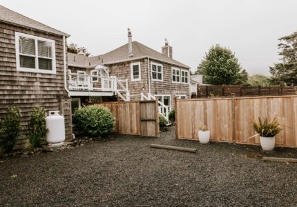 A backyard with dark gravel ground, wooden fences, and a wooden house with shingle siding. The backyard has potted plants, a propane tank, and a staircase leading to a balcony. Trees and greenery are visible in the background.