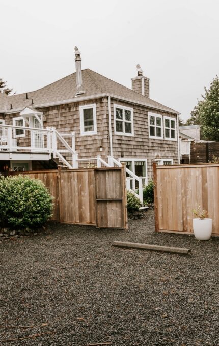 A backyard with dark gravel ground, wooden fences, and a wooden house with shingle siding. The backyard has potted plants, a propane tank, and a staircase leading to a balcony. Trees and greenery are visible in the background.
