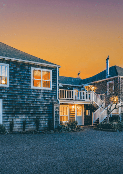 A two-story wooden house with numerous windows and a staircase is lit up from inside during twilight. The exterior features a shingle siding, a fenced area to the right, and a spacious gravel driveway in the foreground. Trees and shrubs surround the house.