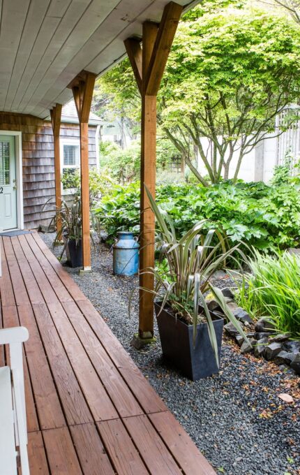 A wooden porch with two white benches extends along the side of a house. Potted plants and a small garden with shrubs and rocks are adjacent to the porch. The porch leads to a white door with glass panels. Trees and foliage surround the area.