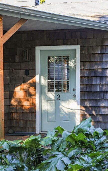 A wooden building with shingle siding and a light green door labeled "2". A small porch area includes a white bench on the left. There are plants and shrubs in the foreground and a window with blinds to the right of the door.