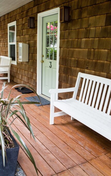 A wooden porch with brown shingle siding and a light green front door. Two white wooden benches are placed near the door, with potted plants and decorative items positioned on the porch. Garden foliage is visible near the porch area.