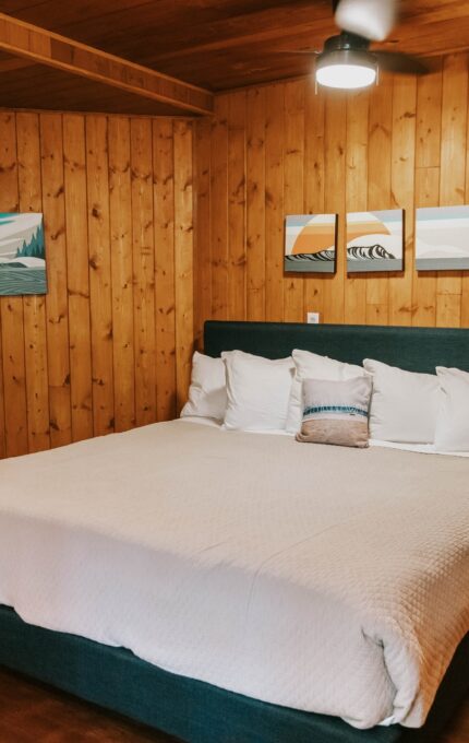 A cozy bedroom features a large bed with a blue upholstered frame and white bedding, flanked by a nightstand with a lamp. The walls and ceiling are covered in wooden panels, and a small armchair is positioned in the corner. Three paintings decorate the wall above the bed.