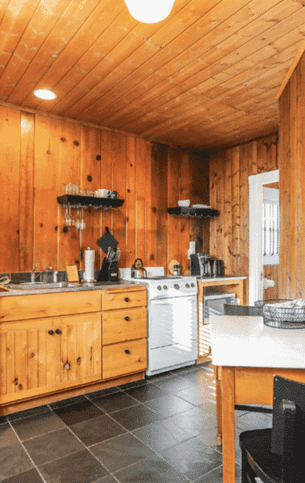 A small kitchen with wooden paneled walls and ceiling. It features a white refrigerator, wooden cabinets, a stove, a sink, and shelves with dishes. A small table with black chairs is in the corner. The floor is tiled in dark gray. Two framed pictures hang on the wall.