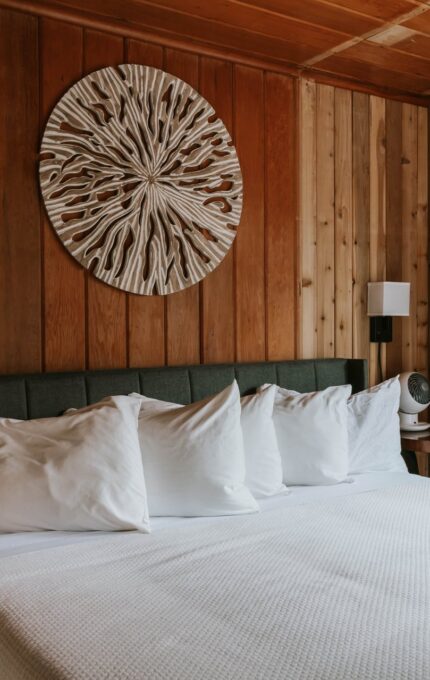 The image shows a cozy bedroom with wooden walls and ceiling. A large bed with a dark headboard features white bedding and multiple pillows. A wooden wall sculpture hangs above the bed. A pair of white bathrobes are hung on the wall near a window.