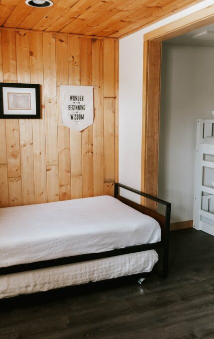 A cozy bedroom featuring a single bed with a trundle, adorned with patterned pillows, and a set of white bunk beds. The walls are made of wooden panels, decorated with framed pictures and a fabric sign reading "Winter Dreaming". The flooring is dark wood.