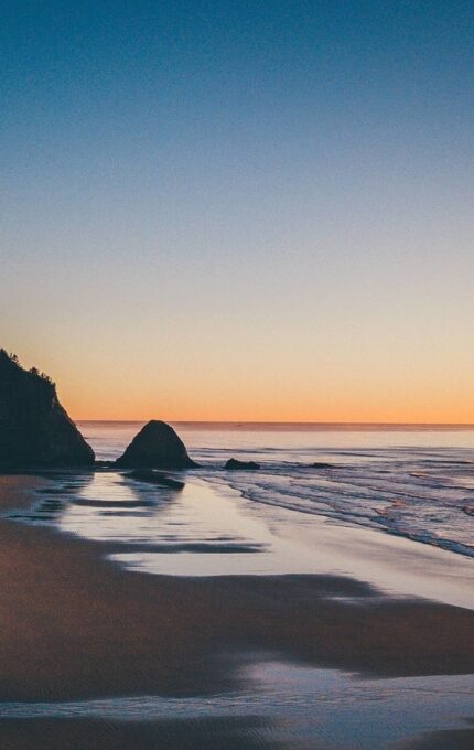 A scenic view of a beach at sunset. The sun is low on the horizon, casting a golden glow over the calm ocean. The shoreline has gentle waves and a large rock near the water's edge. Trees and a few buildings are visible on a hill to the left.