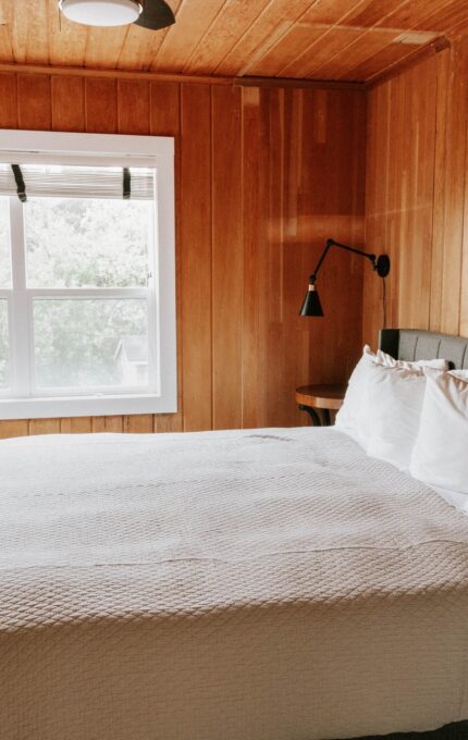 A bedroom with wooden walls and ceiling features a large bed with white bedding and pillows. A black wall-mounted lamp is above the nightstand on the right. A window with blinds partially opened faces the bed, and a chest of drawers is on the left.